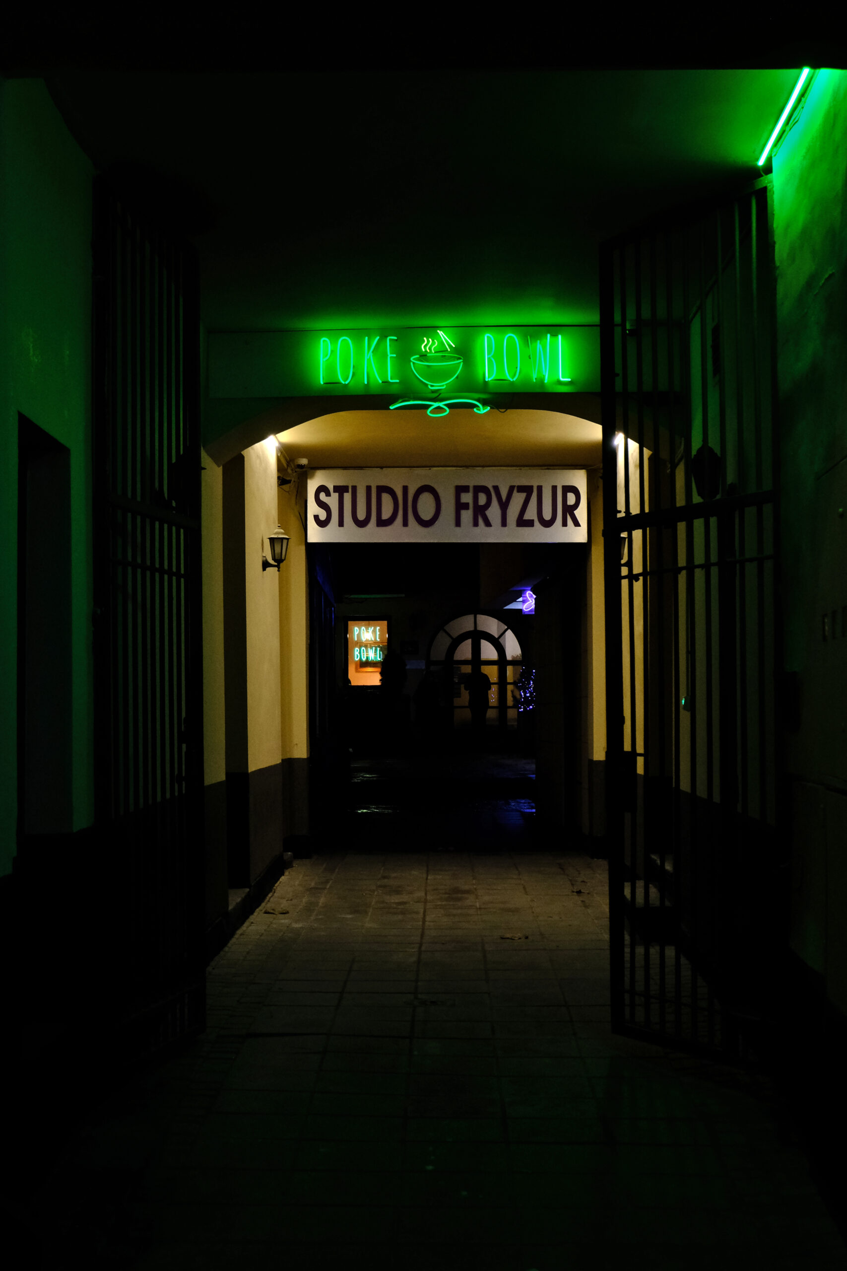 Alley with a green neon sign saying "Poke Bowl". Below and further a sign saying "Studio Fryzur". In the back you can see a small neon sign in the window also saying "Poke Bowl".
