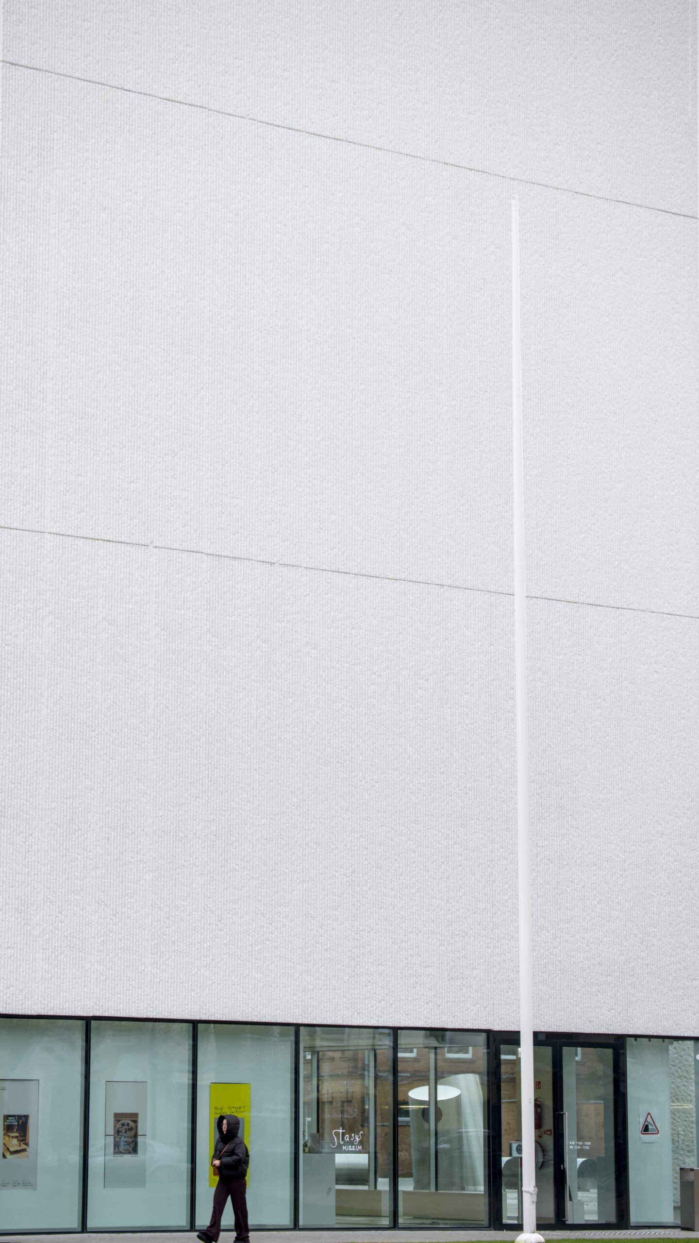 Person walking in front of a tall building with only one wall visible. Corners and top are out of frame. Ground floor is windows, the rest and the majority of the frame is a grooved white wall.