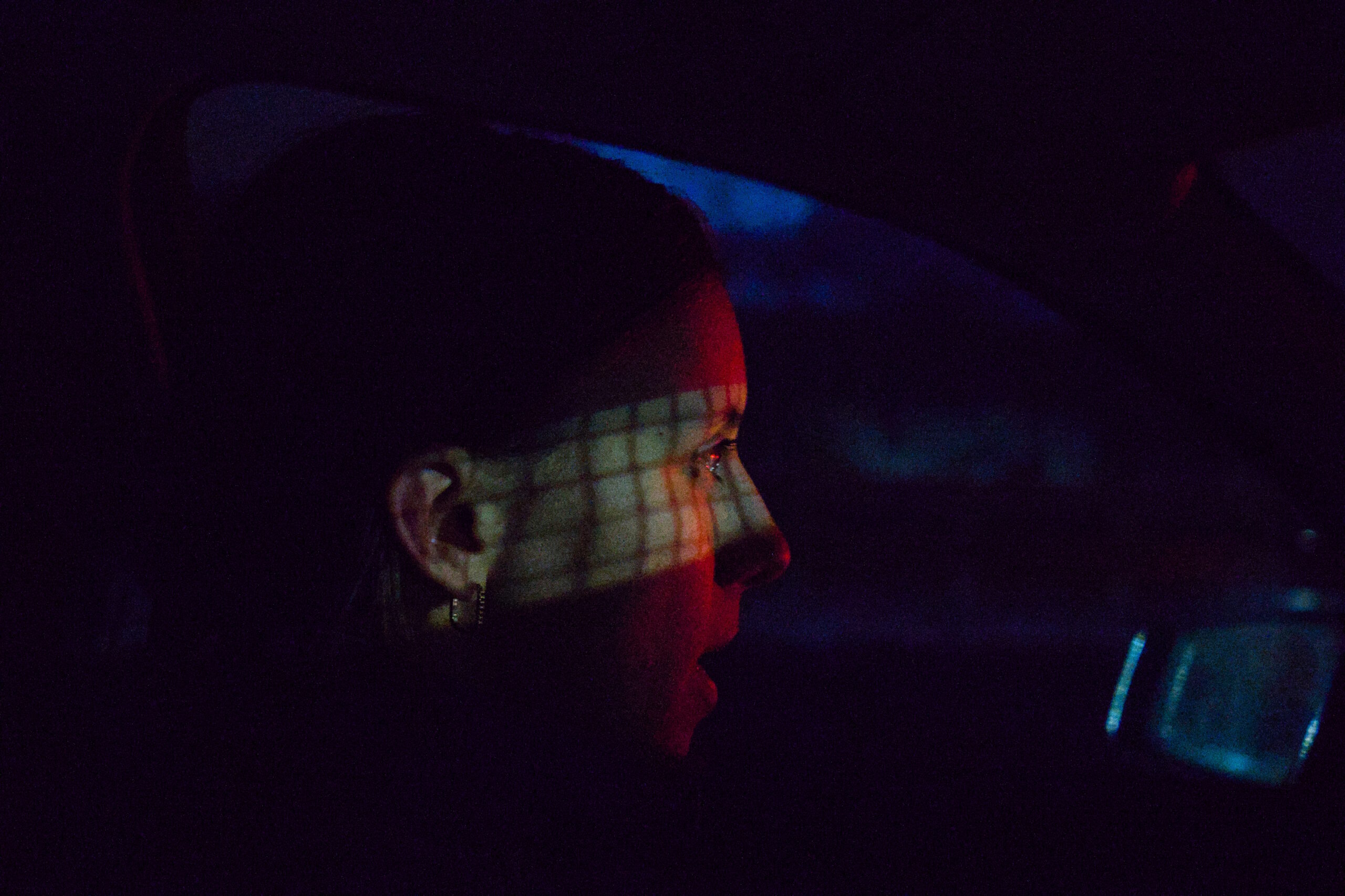Woman driving in the dark, sideview. Red brake light shines onto face from vehicles in front. Rearview mirror reflects white light and grid pattern of a net onto eye and ear.
